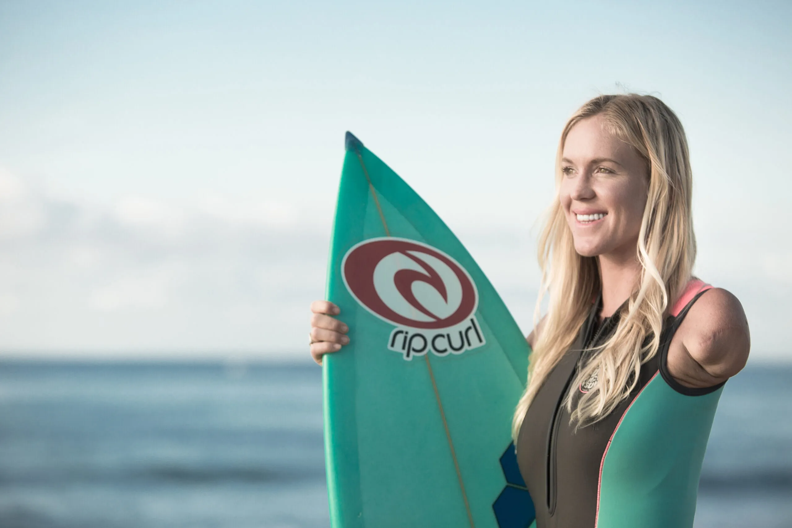Bethany Hamilton, smiling and holding a Rip Curl surfboard with her right arm, stands by the ocean wearing a wetsuit, showcasing her strength and resilience